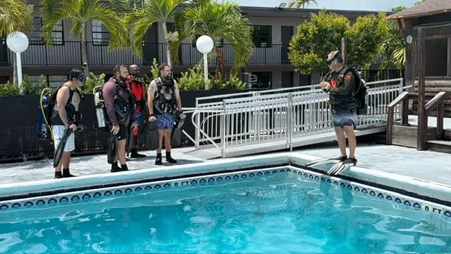Four divers in scuba gear stand at the edge of a pool listening to an instructor