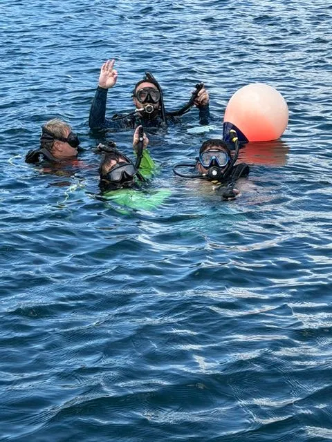 Four divers at the surface of open water next to an orange dive float, signaling OK